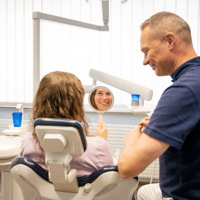 Gesunde-Zaehne-mit-der-Zahnarztpraxis-Dr.-Handschuh-in-Alsfeld-mobil Dr. Christian Handschu berät im Behandlungsraum der Zahnarztpraxis Dr. Handschuh in Alsfeld eine Patientin, die einen Spiegel in der Hand hält und lächelt